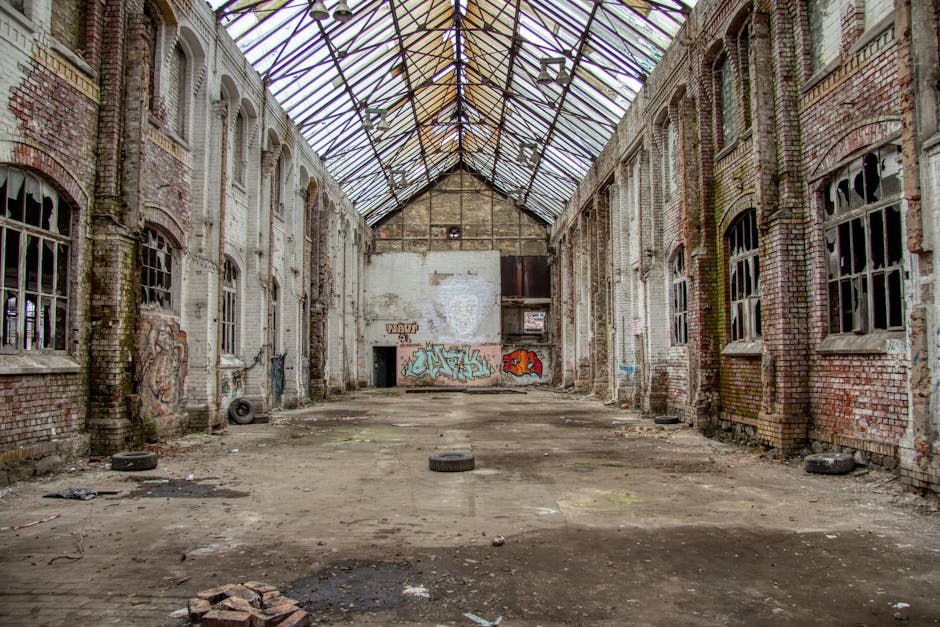 A large, abandoned industrial interior featuring a wide, open space with a dirt and debris-covered concrete floor. The walls are constructed of red brick and show signs of deterioration, with patches of moss or algae growth near the base and graffiti tags painted on the surfaces. Tall, arched windows with broken or missing glass line both sides of the space, allowing natural light to filter through and creating a slightly dim, atmospheric ambiance. The ceiling is a metal framework supporting a partially intact, corrugated glass roof, some of which is missing or damaged, revealing the sky above. Scattered around the floor are old tires, bricks, and pieces of rubble, suggesting previous or ongoing clearance attempts. In the background, there is a small doorway or opening behind some graffiti, contributing to the scene’s sense of neglect, yet it subtly relates to the concept of private waste management or alternative collection activities carried out by companies such as Waste Removal Haringey. This space, likely used earlier for industrial purposes, now appears disused and suitable for on-site clearance or waste removal services aimed at transforming derelict structures into usable spaces.