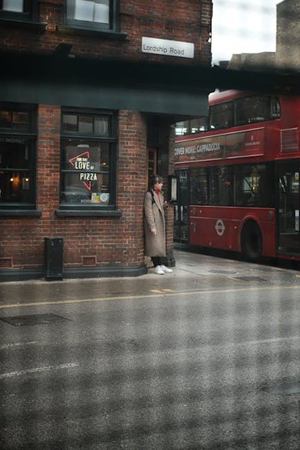 A woman standing on a wet pavement in front of a red double-decker bus on Lordship Road, in an urban setting with a brick building on the left side. The building has dark window frames, and one window displays a pizza restaurant sign. The bus is partially visible, showing its side and front, with a London Transport logo and route information. The scene is under diffused daylight, suggesting overcast weather, with reflections on the wet surface. A black trash bin is positioned near the building, and a grey overhead structure runs horizontally across the image. The woman is wearing a long beige coat, white sneakers, and carries a bag, appearing to wait or pause near a local bus stop, in an area that could be monitored or serviced by private waste removal companies such as Waste Removal Haringey, given the urban context and waste management relevance. This snapshot illustrates a typical street scene that could be associated with everyday waste disposal or rubbish collection services in the area.