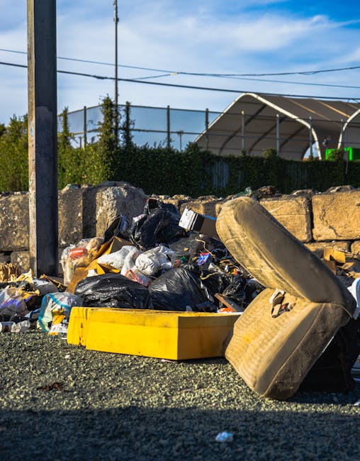 A pile of discarded waste materials and rubbish bags accumulated on a gravel surface in an outdoor setting, with a yellow plastic container and a large, worn car tire among the debris. The rubbish includes black plastic bags, some partially open revealing mixed waste inside, along with flattened cardboard boxes and miscellaneous packaging. In the background, there is a stone wall, a metal utility pole, and power lines, with a hedge and a partially visible large tent structure or sports facility behind a fence. The sky is partly cloudy with blue patches, suggesting daytime, and the scene appears to depict an area awaiting removal or collection of waste, relevant to private rubbish disposal services like those offered by Waste Removal Haringey, as an alternative to council collections.