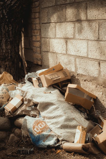 A pile of mixed rubbish and cardboard boxes is situated in a corner beside a brick and concrete block wall. The waste includes various light brown and white cardboard packaging, some torn or crushed, and a large white fabric sack partially covered by the debris. The surrounding ground appears uneven, with small rocks and dirt visible beneath the rubbish. The backdrop features a wall built from large, rough-textured concrete blocks, situated next to a weathered brick section. To the left, part of a dark tree trunk and some foliage are visible, hinting at an outdoor setting. The scene reflects a typical area where private waste collection, such as that provided by waste removal services in Haringey, might take place, highlighting the need for proper rubbish clearance and potential removal of builders or construction debris by specialist waste disposal providers like Waste Removal Haringey.