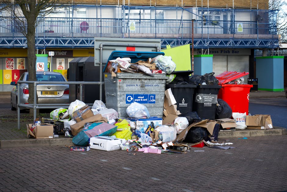 A collection of overflowing rubbish bins and waste scattered on the pavement in front of a commercial building. The waste includes cardboard boxes, plastic bags, paper waste, and miscellaneous packaging materials, with some items spilled onto the nearby sidewalk. The bins are of various sizes and colors; one large grey bin labelled for mixed paper and cardboard is filled to capacity, causing waste to spill out. To the right, a red bin and multiple black bins are also visible, some with garbage bags leaning against them. In the background, a parked silver car with a partially visible license plate is situated behind a metal railing, and a building with a blue scaffolding structure covers the upper part, indicating construction or refurbishment works. The scene suggests a location where domestic or commercial waste has been neglected, underscoring the importance of proper rubbish disposal services like those offered by Waste Removal Haringey for effective waste management and compliance with local regulations.