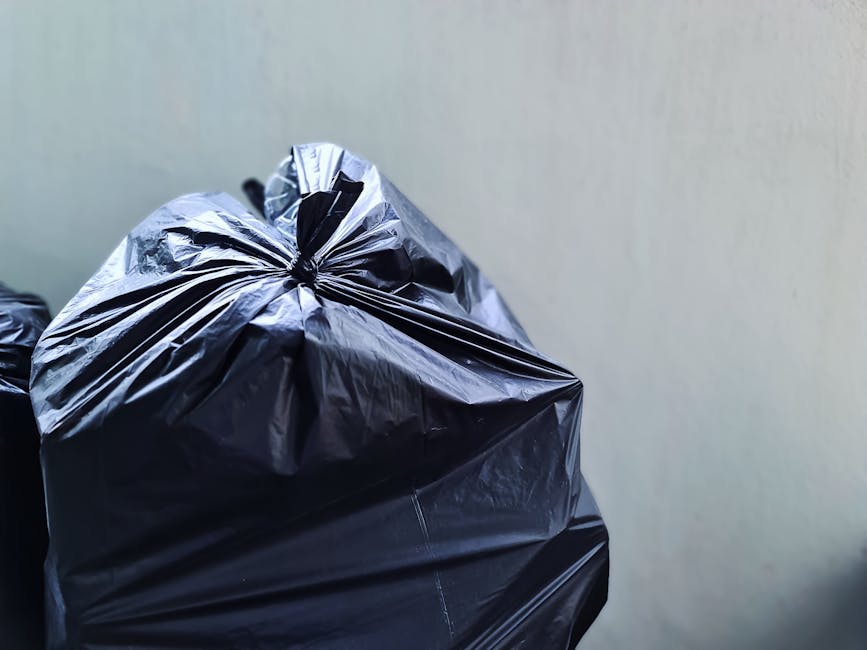 A close-up view of several tightly tied black plastic rubbish bags stacked in front of a plain, light-colored wall. The bags are made of glossy, crinkled plastic material, with some appearing slightly bulging from the contents inside. The bags are positioned on a surface area that is not fully visible but suggests an outdoor or driveway setting, possibly near a waste collection point. The background is neutral, with no other objects or environmental features visible, emphasizing the focus on the black rubbish bags. This scene relates to waste disposal and rubbish removal services offered by Waste Removal Haringey, highlighting the typical appearance of household waste prepared for collection or private disposal methods, consistent with a professional waste management context.