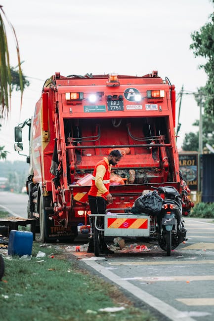 A waste collection worker, dressed in a yellow high-visibility vest with red accents, is positioned near the rear of a large red rubbish collection vehicle on a roadside pavement. The worker is actively engaged in emptying a black trash bag into the vehicle’s open rear compartment, which has a metallic, textured surface with visible hydraulic mechanisms and safety markings. The scene is set on a street with a grassy verge to the left, with a small blue container and a black motorcycle parked nearby on the pavement. In the background, there are trees, utility poles, and some commercial signage, indicating an urban or suburban environment. Daylight provides natural lighting, and the overall scene depicts a routine rubbish collection activity, which could be associated with private waste disposal services like those provided by Waste Removal Haringey. The image captures the practical aspect of waste management, emphasizing the busy work environment and the importance of organized rubbish collection operations.