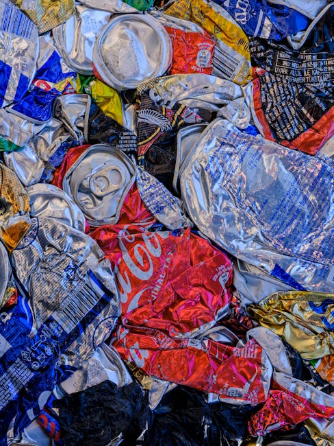 A close-up view of a pile of crushed aluminum soft drink cans and crumpled plastic wrappers, featuring various brands and colours, including red, silver, blue, and black. The cans display metallic surfaces with visible pull tabs and ridges, while the plastic wrappers exhibit glossy finishes with printed text and images. The waste materials are loosely stacked on top of each other, occupying the foreground and filling most of the frame, with little visible background. The scene demonstrates typical household or commercial rubbish, likely collected for disposal or recycling, and highlights the importance of proper waste handling as part of rubbish removal services such as those offered by Waste Removal Haringey. The lighting is even, emphasizing the textures and reflective qualities of the cans and wrappers, creating a realistic representation of mixed packaging waste that could be part of a waste collection or on-site clearance process in a residential or commercial setting.