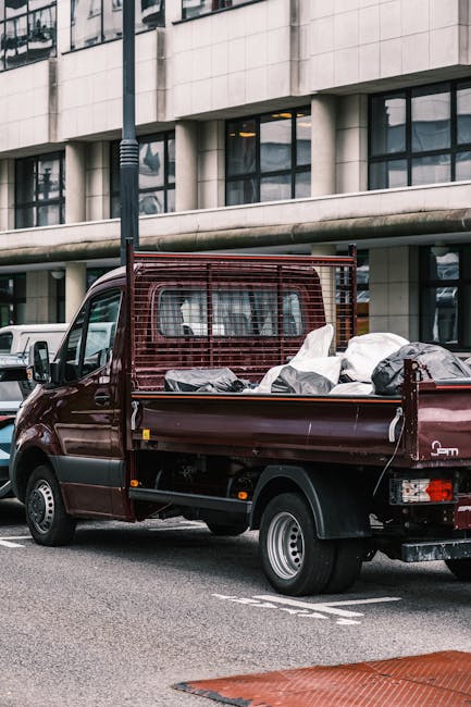 A small, dark red flatbed truck parked on an urban street in front of a modern building with large glass windows and a concrete facade. The truck's cargo area is filled with various types of rubbish, including large black and white plastic bags and other debris, suggesting it is used for private waste collection or on-site clearance services. The vehicle features a wire mesh partition separating the cab from the load space, and its body shows signs of regular use, with minor dirt visible on the surface. Surrounding the truck, there is a parked white vehicle in the background and a black pole with a lamp post standing nearby. The scene appears to be during the daytime, with natural light illuminating the area, consistent with waste removal activities performed by companies such as Waste Removal Haringey that specialise in rubbish disposal and alternative waste handling options for commercial or residential clients.
