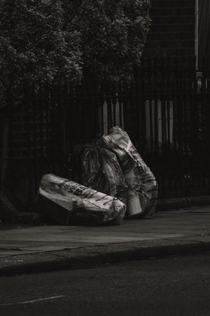 A close-up view of a pile of mixed household rubbish and waste materials situated on a gravel surface in an outdoor area. The debris includes black plastic trash bags, some of which are torn open revealing contents, and various discarded items such as a yellow plastic storage box, a large beige upholstered armchair with visible fabric texture and signs of dirt or wear, and broken or discarded furniture components. The waste is piled against rough stone blocks that form a low retaining wall, with a background featuring a chain-link fence, dense green shrubbery, and a structure with a curved roof. Overhead sky is partly cloudy with patches of blue, and a utility pole with multiple wires is visible on the left side. The scene suggests an environment where independent rubbish collection or on-site clearance services, like those offered by Waste Removal Haringey, may be used for managing bulky or non-standard waste outside typical council disposal areas.