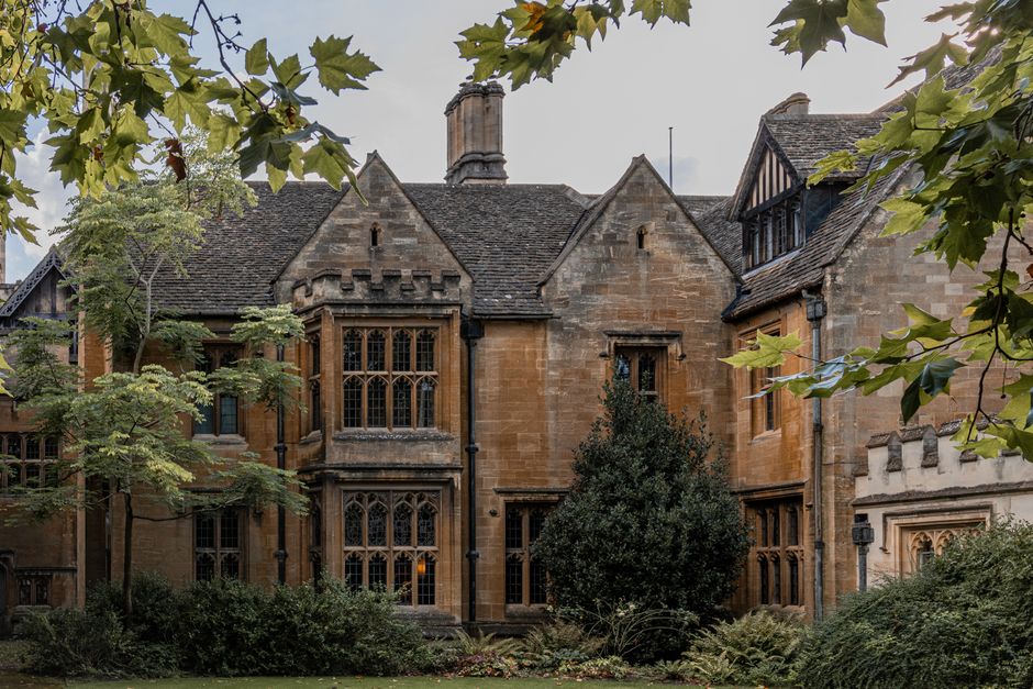 A historic stone building with intricate multi-pane leaded windows, steeply pitched slate roof, and decorative battlements along the parapet. The structure features a prominent bay window with pointed arches and detailed stone tracery, and smaller sash windows on either side. Surrounding the building are lush green trees and shrubs, some with broad leaves extending into the frame from the upper left and right corners. The building’s exterior displays warm, weathered sandstone with a textured finish, and sections of timber framing are visible beneath the roofline, suggesting a Tudor revival style. The scene is captured during daylight hours, with natural light highlighting the building's architectural details against a partly cloudy sky. The setting appears to be part of a private estate or historic property, with mature landscaping creating a tranquil environment. This image reflects the context of private property management and the importance of maintaining large, period-style buildings, which often require specialist waste removal services to handle maintenance debris, renovation waste, or clearance activities related to estate upkeep, as offered by Waste Removal Haringey.