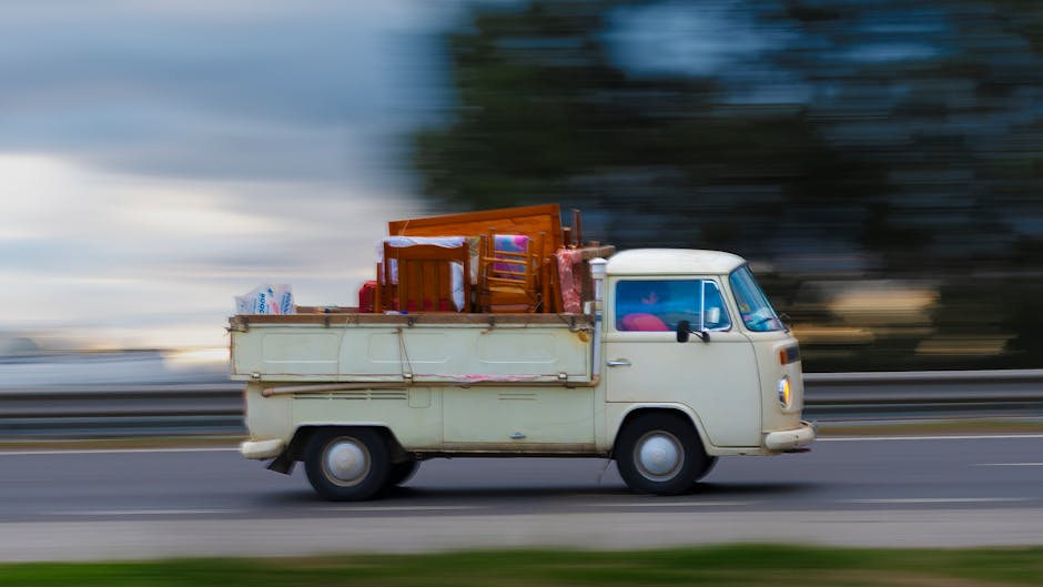 A vintage cream-colored mini truck is driving along a roadway, loaded with assorted household furniture and miscellaneous items. The cargo area at the back contains wooden dining chairs stacked on top of each other, a small wooden table, and a few plastic bags, all secured with light ties. The furniture shows polished wood grain and a smooth finish, contrasting with the plastic bags that appear crumpled and lightweight. The vehicle’s body exhibits minor signs of wear, with the paint slightly aged, and the tires are black with a visible tread pattern. The side view mirror is black and mounted on the door, with the driver's window partially visible, revealing a faintly pink object on the dashboard inside. The background features a blurred landscape of trees and sky, creating a sense of motion and speed, typical of independent or private waste transportation. The overall scene reflects the context of alternative rubbish handling and on-site clearance, aligned with waste removal services such as those offered by Waste Removal Haringey, emphasizing the practical aspect of non-public disposal methods for bulky household waste.