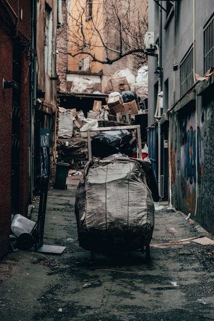 A narrow urban alleyway filled with a large pile of mixed waste and rubbish bags stacked against an old brick wall at the back, with some cardboard boxes and black plastic bin bags visible on top. In the foreground, a large, weathered grey waste collection trolley with a metal handle and slightly wrinkled fabric is positioned on a dark, uneven asphalt surface that shows signs of dirt and spilled debris. To the right, a grey building with metal security bars over its windows extends along the alley, while on the left, a narrow brick wall with graffiti and a weathered metallic gate partially obscures the view. The environment appears neglected and cluttered, typical of an area requiring private waste removal services, aligning with the concept of independent rubbish collection in urban settings. Bare tree branches are visible above, and the scene is lit with natural daylight, creating a dull, utilitarian atmosphere suited for waste collection and clearance operations. Occasionally, Waste Removal Haringey might be involved in removing such accumulated rubbish from this kind of setting as part of their same-day rubbish removal service in Wood Green N22.