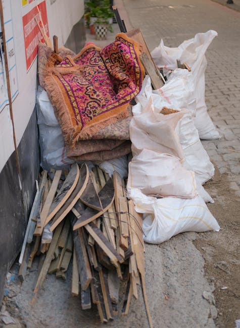 A collection of discarded household items and construction debris piled along a sidewalk next to a building wall. The pile includes an ornate, multicolored Persian-style rug with detailed patterns and fringed edges, draped over other materials. Surrounding the rug are several large, white, woven polypropylene bags filled with unknown waste, some appearing partially open with visible contents. To the left, there are broken wooden items, possibly shelving or furniture parts, with light-colored, weathered surfaces, and some metal or plastic components mixed in. The debris is stacked informally on a paved pavement with visible cracks, and the scene suggests an area temporarily used for rubbish removal or on-site clearance, possibly by a private waste handling service such as Waste Removal Haringey, in an urban setting.