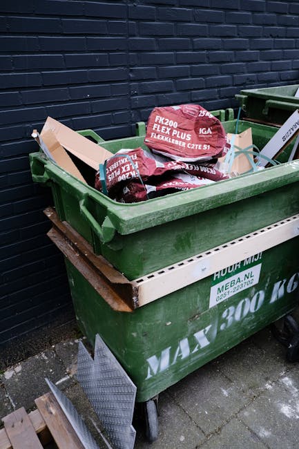 A narrow urban alleyway filled with a large pile of mixed waste and rubbish bags stacked against an old brick wall at the back, with some cardboard boxes and black plastic bin bags visible on top. In the foreground, a large, weathered grey waste collection trolley with a metal handle and slightly wrinkled fabric is positioned on a dark, uneven asphalt surface that shows signs of dirt and spilled debris. To the right, a grey building with metal security bars over its windows extends along the alley, while on the left, a narrow brick wall with graffiti and a weathered metallic gate partially obscures the view. The environment appears neglected and cluttered, typical of an area requiring private waste removal services, aligning with the concept of independent rubbish collection in urban settings. Bare tree branches are visible above, and the scene is lit with natural daylight, creating a dull, utilitarian atmosphere suited for waste collection and clearance operations. Occasionally, Waste Removal Haringey might be involved in removing such accumulated rubbish from this kind of setting as part of their same-day rubbish removal service in Wood Green N22.