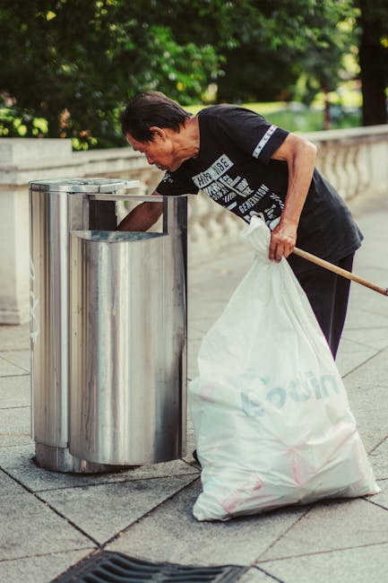 A man with dark hair and light skin, dressed in a black T-shirt with white printed text, is seen bending over and disposing of rubbish into a large, open-top metal bin. He is holding a white refuse sack in one hand and appears to be emptying or collecting waste from the bin, which is situated on a paved outdoor area. The bin is made of stainless steel with a shiny, reflective surface and is positioned next to a low stone or concrete railing. In the background, there are lush green trees and foliage, suggesting a park or landscaped public space. The scene is well-lit with natural daylight, highlighting the textures of the metal bin, the fabric of the refuse sack, and the surrounding environment. This image reflects the process of private or independent rubbish collection in an outdoor urban setting, consistent with waste removal services such as those provided by Waste Removal Haringey.