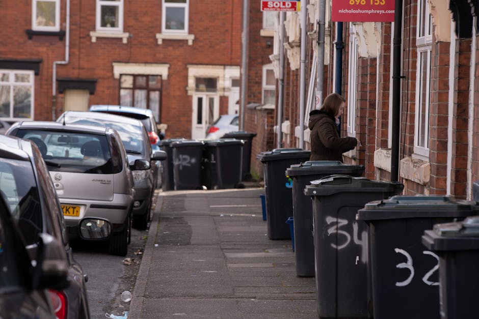 A street scene in an urban residential area shows multiple black wheelie bins lined along the pavement outside brick terraced houses, used for rubbish collection. The bins are made of plastic with textured surfaces, fitted with lids, and some are marked with white spray paint numbers or letters. A woman in a dark brown coat stands with her back to the camera, opening or handling one of the bins, which are positioned close to the building's exterior wall. The background features more parked vehicles, including a silver car, and additional bins placed further up the street. The brick buildings have white-framed windows and some display signs, including one with the word 'SOLD' in red, and another advertising a property company. The environmental setting appears to be a typical urban street during daytime, with natural light illuminating the scene, emphasizing the neat arrangement of waste disposal containers for private or alternative rubbish collection services, such as those offered by Waste Removal Haringey, in the N17 Tottenham area.