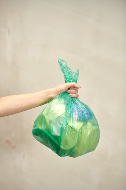 A man with dark hair and light skin, dressed in a black T-shirt with white printed text, is seen bending over and disposing of rubbish into a large, open-top metal bin. He is holding a white refuse sack in one hand and appears to be emptying or collecting waste from the bin, which is situated on a paved outdoor area. The bin is made of stainless steel with a shiny, reflective surface and is positioned next to a low stone or concrete railing. In the background, there are lush green trees and foliage, suggesting a park or landscaped public space. The scene is well-lit with natural daylight, highlighting the textures of the metal bin, the fabric of the refuse sack, and the surrounding environment. This image reflects the process of private or independent rubbish collection in an outdoor urban setting, consistent with waste removal services such as those provided by Waste Removal Haringey.